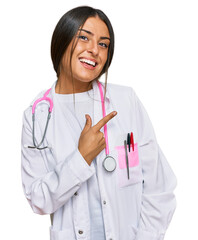Beautiful hispanic woman wearing doctor uniform and stethoscope cheerful with a smile of face pointing with hand and finger up to the side with happy and natural expression on face