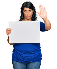 Plus size brunette woman holding blank empty banner pointing with finger to the camera and to you, confident gesture looking serious
