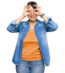 Young beautiful hispanic woman with short hair wearing casual denim jacket doing ok gesture like binoculars sticking tongue out, eyes looking through fingers. crazy expression.