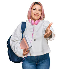 Hispanic woman with pink hair wearing student backpack and headphones smiling cheerful offering...