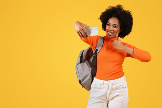 Smiling young woman with afro hair taking a selfie and giving a thumbs up, wearing an orange turtleneck