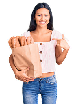 Young beautiful latin girl holding paper bag with bread pointing finger to one self smiling happy and proud