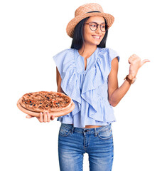 Obraz premium Young beautiful latin girl wearing summer hat and glasses holding pizza pointing thumb up to the side smiling happy with open mouth
