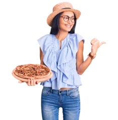 Poster Im Rahmen Young beautiful latin girl wearing summer hat and glasses holding pizza pointing thumb up to the side smiling happy with open mouth © Krakenimages.com
