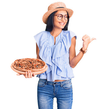 Fototapeta Young beautiful latin girl wearing summer hat and glasses holding pizza pointing thumb up to the side smiling happy with open mouth
