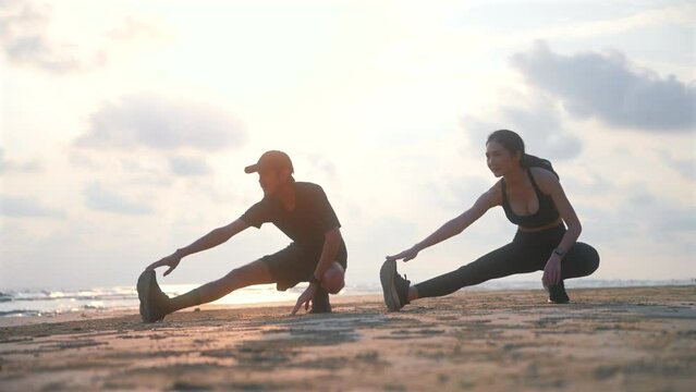 Happy asian sports couple stretching exercises at seashore with dramatic sky and bright sunset. Silhouette of man and woman jogger during outdoor activity on beach. 4k resolution and slow motion shot.