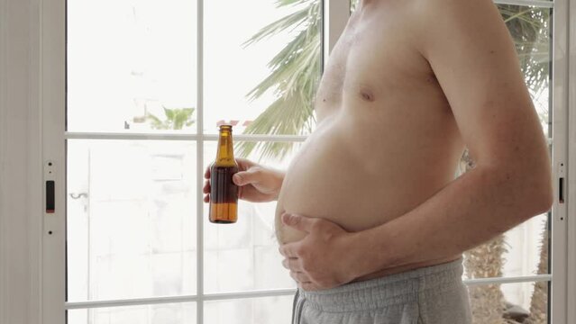 man with  beer belly stands on terrace, sipping beer,Patting his chubby belly