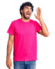 Handsome young man with curly hair and bear wearing casual pink tshirt shouting and screaming loud to side with hand on mouth. communication concept.