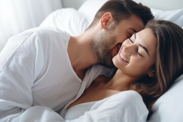 Young affectionate couple smiling and cuddling in a white bed, showing happiness and love. Intimate Couple Smiling in Bed Together - obrazy, fototapety, plakaty