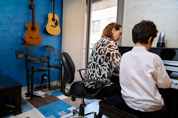 Rear view of a teenage student boy playing piano, composing a melody under his teacher's guidance...