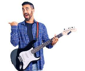 Young hispanic man playing electric guitar pointing thumb up to the side smiling happy with open mouth