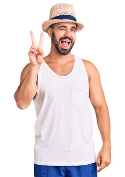 Young hispanic man wearing casual summer hat smiling with happy face winking at the camera doing victory sign. number two.