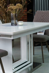 White wooden table combined with gray wooden chairs with soft fabric upholstery. There is a black glass vase with dried flowers on the table