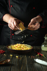 A cook grates hard cheese into a plate of pasta with a hand grater. Working environment in restaurant kitchen with spices and kitchen utensils