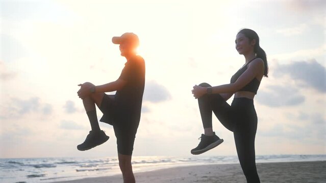 Happy asian sports couple stretching exercises at seashore with dramatic sky and bright sunset. Silhouette of man and woman jogger during outdoor activity on beach. 4k resolution and slow motion shot.