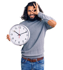 Young arab man holding big clock smiling happy doing ok sign with hand on eye looking through fingers
