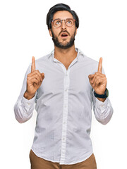 Young hispanic man wearing business shirt and glasses amazed and surprised looking up and pointing with fingers and raised arms.