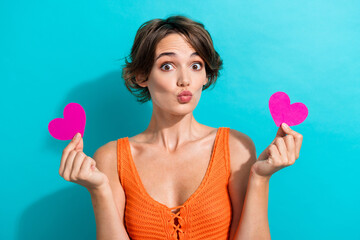 Portrait of coquettish girl with bob hairstyle wear orange knit top hold hearts postcards plump lips isolated on blue color background