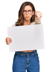 Young brunette woman holding blank empty banner pointing with finger to the camera and to you, confident gesture looking serious