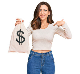 Young brunette woman holding dollars bag pointing finger to one self smiling happy and proud