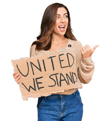 Young brunette woman holding united we stand banner pointing thumb up to the side smiling happy with open mouth
