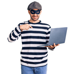 Young handsome man wearing burglar mask using laptop pointing finger to one self smiling happy and proud