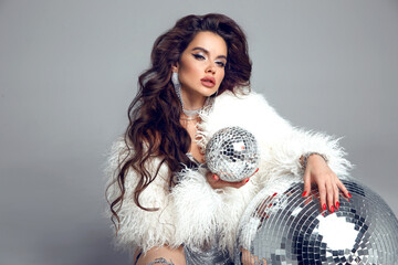 Disco girl. Glamorous brunette in sexy dress with white fur coat over disco glass balls. New Year party. Nightclub. Beautiful lady posing isolated on studio background.