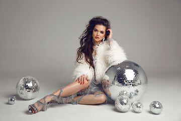 Disco girl. Glamorous brunette in sexy dress with white fur coat over disco glass balls. New Year party. Nightclub. Beautiful lady posing long legs isolated on studio background.