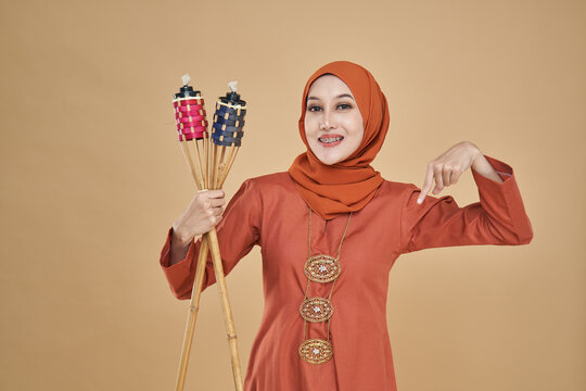 Beautiful Asian model wearing orange "baju kurung" dress with hijab, holding a bamboo torch over beige background.  Eidul fitri festival, fashion and beauty concept.