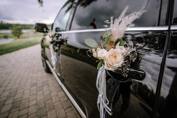 A floral decoration with white roses and feathers is attached to the door handle of a black car,...