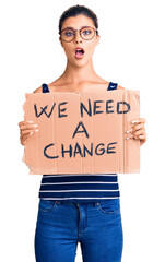 Young beautiful woman holding we need a change banner scared and amazed with open mouth for...