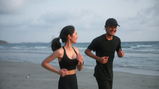 Happy asian sports couple running at seashore with dramatic sky and bright sunset. Silhouette of man and woman jogger during outdoor activity on beach. 4k resolution and slow motion shot.