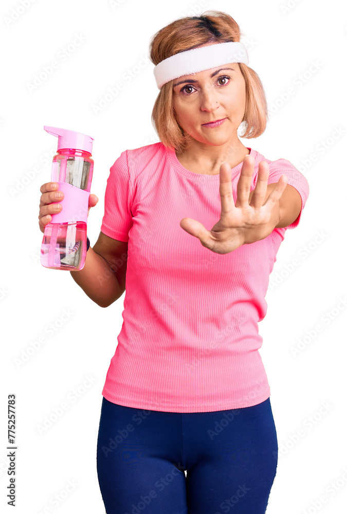 Sticker young blonde woman wearing sportswear holding water bottle with open hand doing stop sign with serio