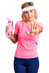Young blonde woman wearing sportswear holding water bottle with open hand doing stop sign with serious and confident expression, defense gesture