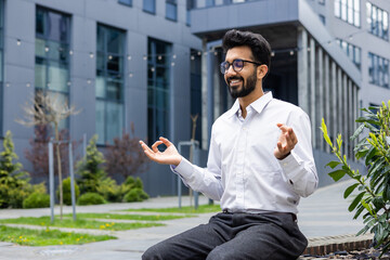 Smiling and relaxed young Indian male businessman sitting on bench in lotus position near offices,...