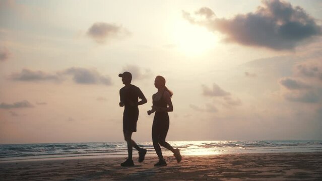 Happy asian sports couple running at seashore with dramatic sky and bright sunset. Silhouette of man and woman jogger during outdoor activity on beach. 4k resolution and slow motion shot.