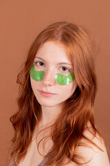 Portrait of a young woman with red hair and green under-eye patches on a beige background, looking at the camera.