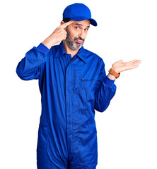 Middle age handsome man wearing mechanic uniform confused and annoyed with open palm showing copy space and pointing finger to forehead. think about it.