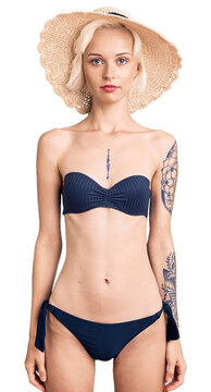 Young blonde woman with tattoo wearing bikini and summer hat relaxed with serious expression on face. simple and natural looking at the camera.