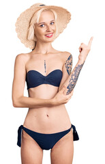 Young blonde woman with tattoo wearing bikini and summer hat with a big smile on face, pointing with hand and finger to the side looking at the camera.