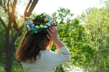 Girl in decorative Spring wreath in garden, natural background. rear view. Floral crown with eggs, symbol of Easter, Ostara holiday. festive decor for spring season. ceremony for wiccan Ostara sabbat - obrazy, fototapety, plakaty