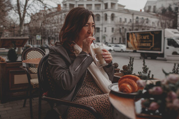 A charmig middle-aged woman drinks latte and eats  croissant in a french cafe on a city street. A...