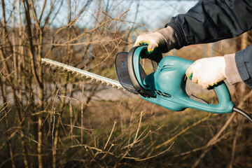 Close-up of a man using an electric brush cutter to cut off the branches in the garden on a spring day. - obrazy, fototapety, plakaty