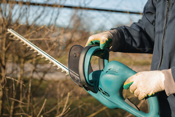 Close-up of a man using an electric brush cutter to cut off the branches of a bush. Springtime. - obrazy, fototapety, plakaty