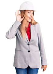 Beautiful young woman wearing architect hardhat smiling with hand over ear listening an hearing to rumor or gossip. deafness concept.