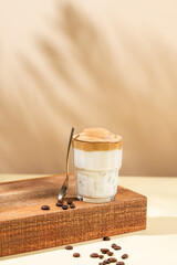 Dalgona coffee. Whipped instant coffee in a glass on a wooden stand with coffee beans on a beige...