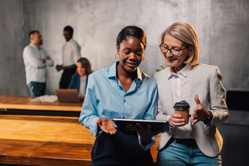Interracial businesswomen discussing report on tablet at workspace.