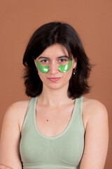A brunette girl wearing green eye patches and a matching sports top looks directly at the camera against a brown background. Her expression exudes calmness and confidence.