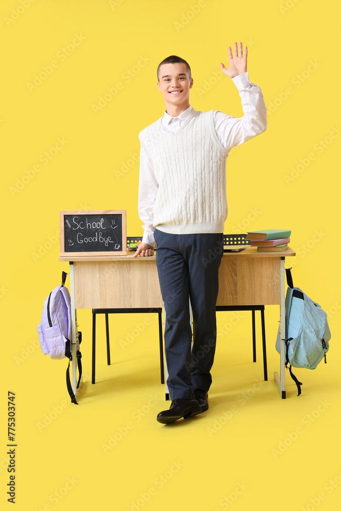 Wall mural Male student near desks with chalkboard with text GOODBYE SCHOOL waving hand on yellow background. End of school concept