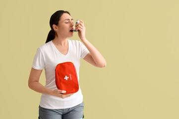 Sporty young woman with first aid kit using inhaler on green background